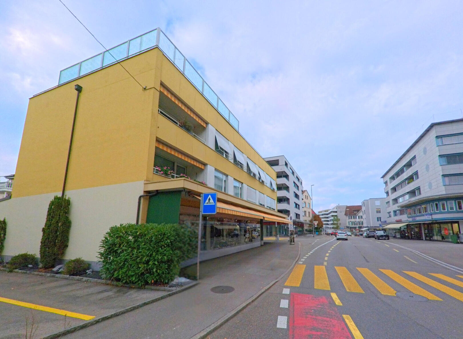 In Wohlen (5610, Kanton Aargau) verläuft eine Strasse mit modernen Wohn- und Geschäftshäusern, an der ein Fussgänger den Zebrastreifen überquert.