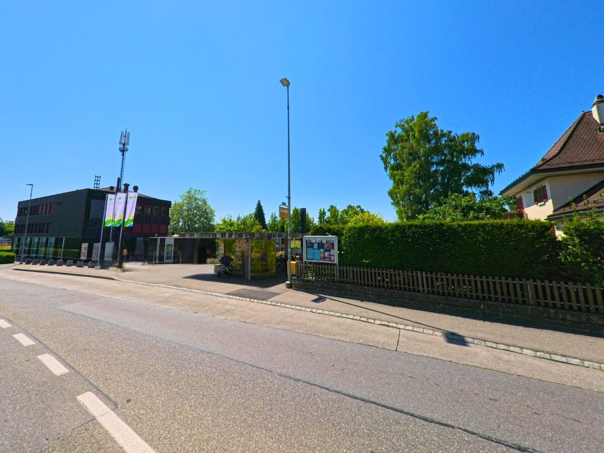 In Wohlen bei Bern (3033, Kanton Bern) befindet sich eine moderne Bushaltestelle mit überdachtem Wartebereich gegenüber einem Verwaltungsgebäude und neben einem Wohnhaus mit gepflegter Hecke.