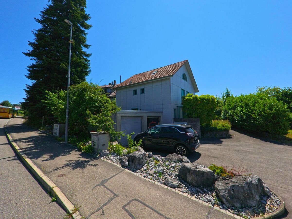 In einer Wohnstrasse von Wohlen bei Bern (3033, Kanton Bern) steht ein modernes Einfamilienhaus mit Garage und Kiesbeet, während im Hintergrund ein gelber Postbus die Anhöhe passiert.