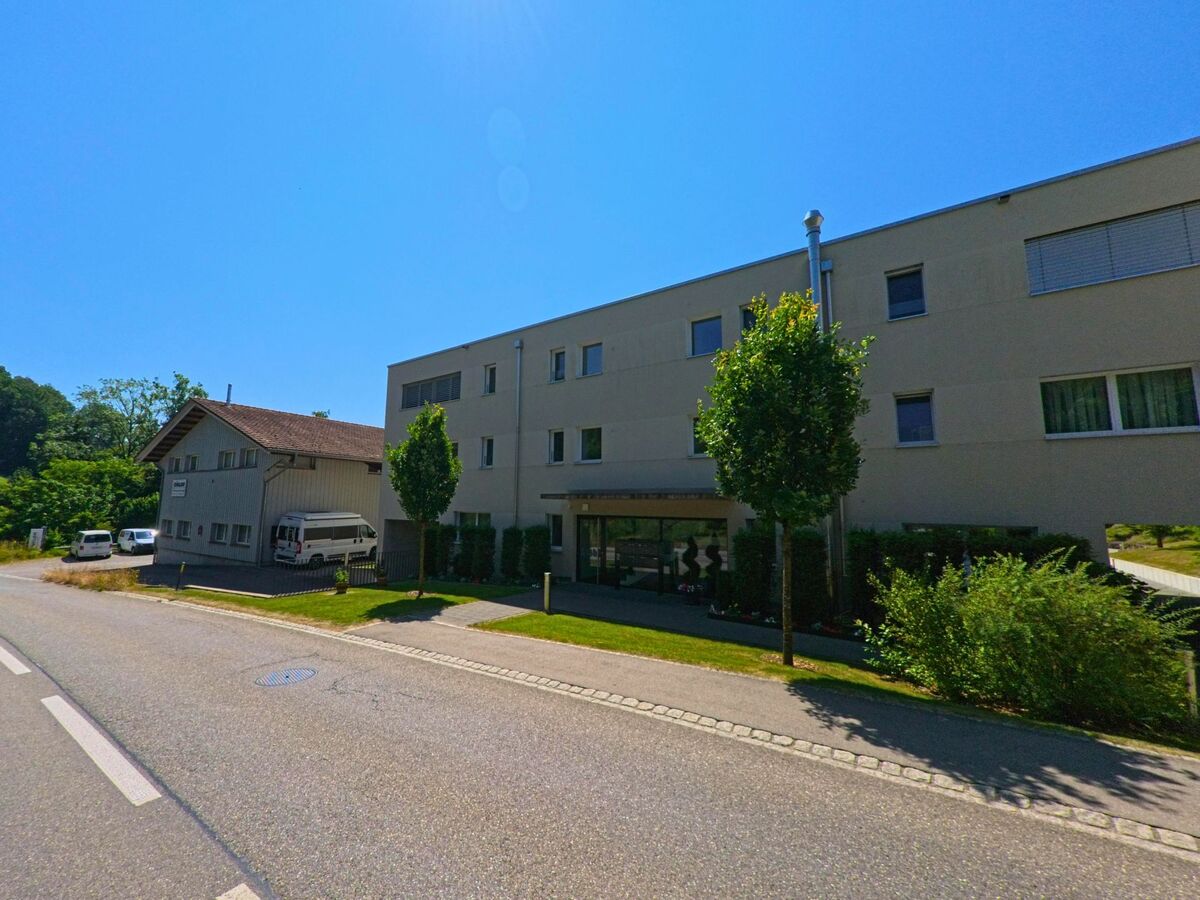 In Wohlen bei Bern (3033, Kanton Bern) steht ein modernes Flachdachgebäude mit gepflegtem Eingangsbereich neben einem älteren Gewerbebau mit Parkplätzen und Lieferwagen.