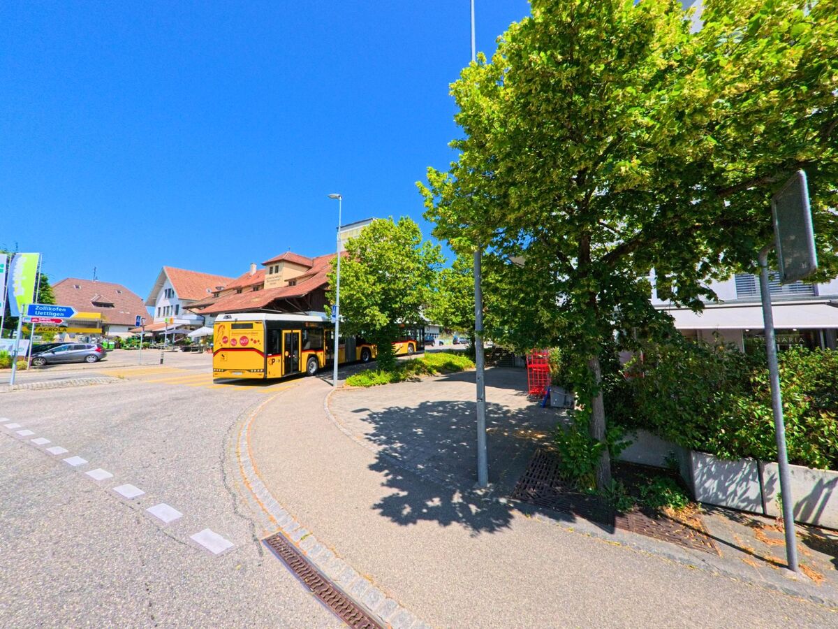 Im Zentrum von Wohlen bei Bern (3033, Kanton Bern) halten zwei gelbe Postbusse an einer belebten Strassenkreuzung mit angrenzenden Restaurants, Geschäften und Wohnhäusern.