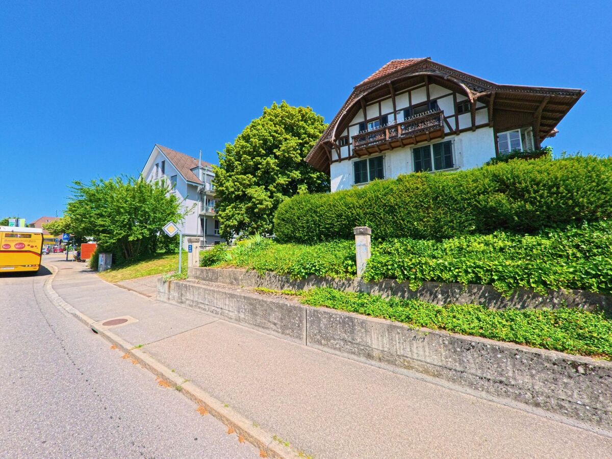 In Wohlen bei Bern (3033, Kanton Bern) verläuft eine leicht gebogene Strasse an einem traditionellen Wohnhaus mit markantem Holzbalkon und gepflegter Hecke vorbei, während ein gelber Postbus die Kurve nimmt.
