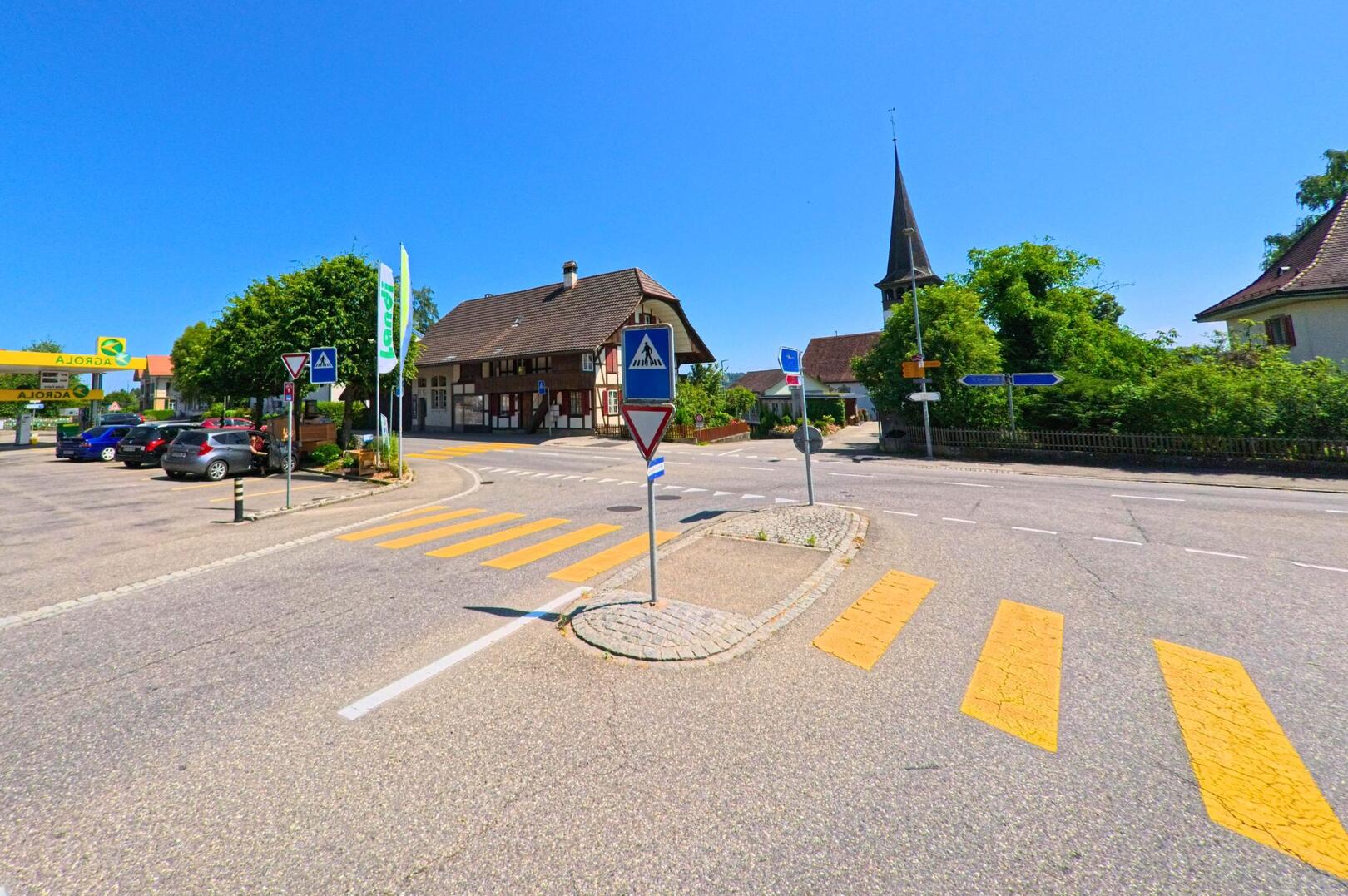 Im Zentrum von Wohlen bei Bern (3033, Kanton Bern) treffen sich mehrere Strassen an einer Verkehrsinsel, flankiert von einem Fachwerkhaus, einer Tankstelle und dem Kirchturm des Dorfes.