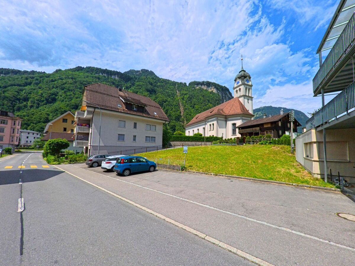 Die Kirche von Wolfenschiessen (6386, Kanton Nidwalden) steht am Fuss eines bewaldeten Hangs, umgeben von Wohngebaeuden und einer stillen Nebenstrasse mit Parkplaetzen.