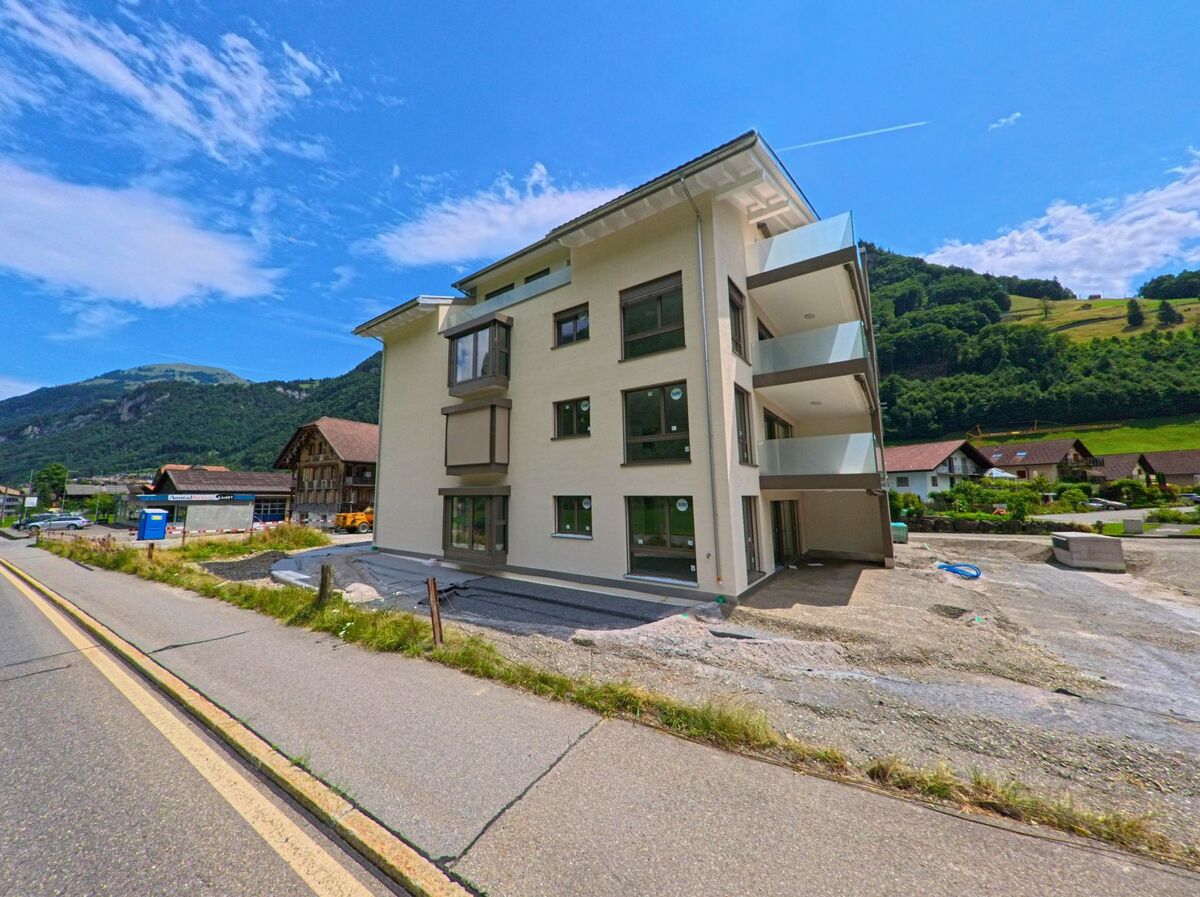 In Wolfenschiessen (6386, Kanton Nidwalden) steht ein neu erbautes Mehrfamilienhaus mit Balkonen an einer Hauptstrasse, umgeben von traditionellen Bauten und gruenen Haengen.