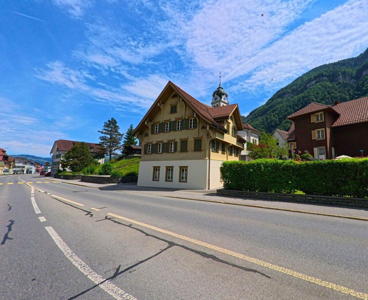 Ein traditionelles Wohnhaus mit gruenen Fensterlaeden steht in Wolfenschiessen (6386, Kanton Nidwalden) an einer Hauptstrasse, im Hintergrund erhebt sich der markante Kirchturm vor bewaldetem Hang.