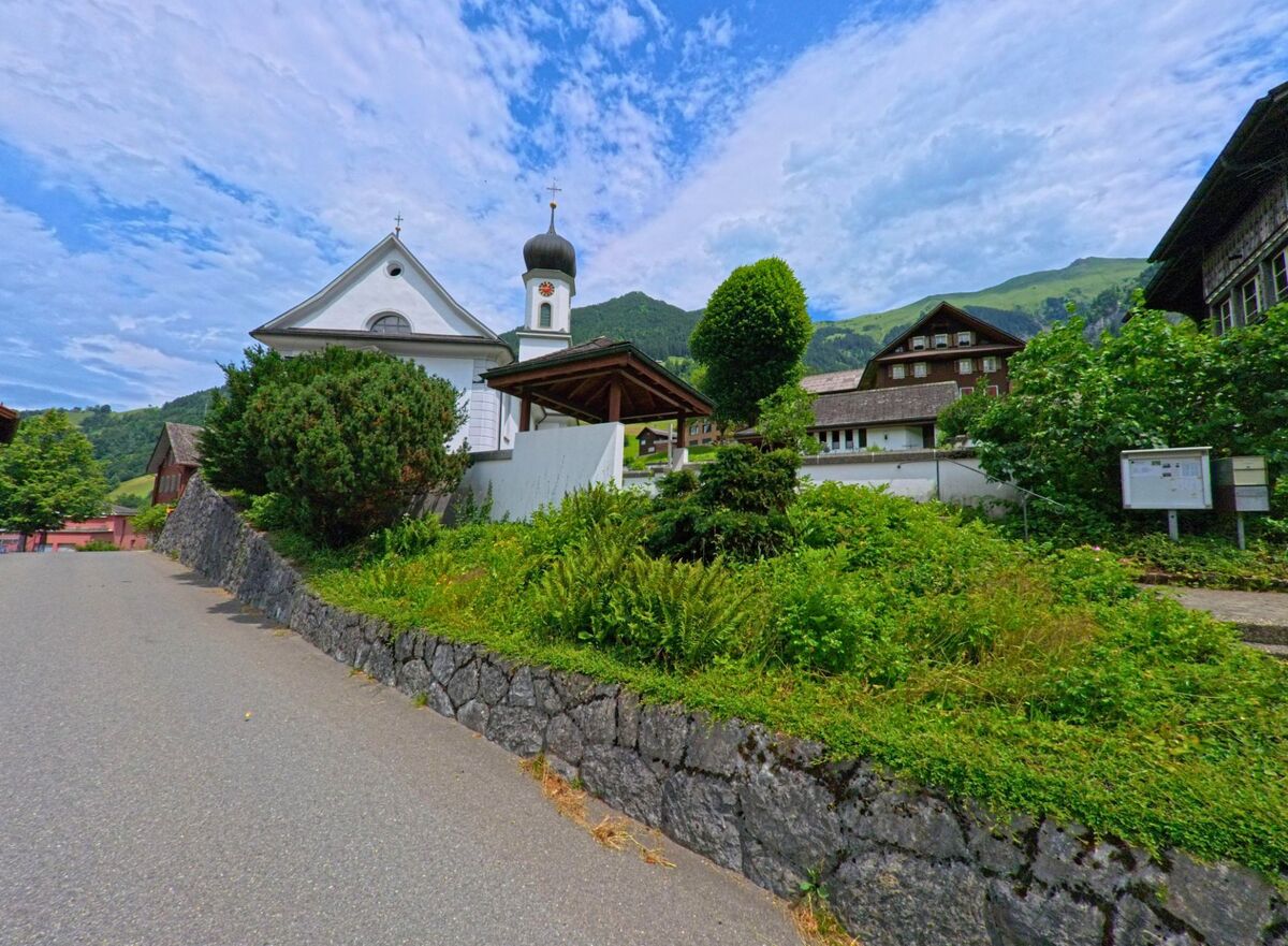 In Wolfenschiessen (6387, Kanton Nidwalden) liegt eine kleine Kirche mit Zwiebelturm am Hang, umgeben von traditionellen Holzhäusern und dichter Vegetation vor einer alpinen Bergkulisse.