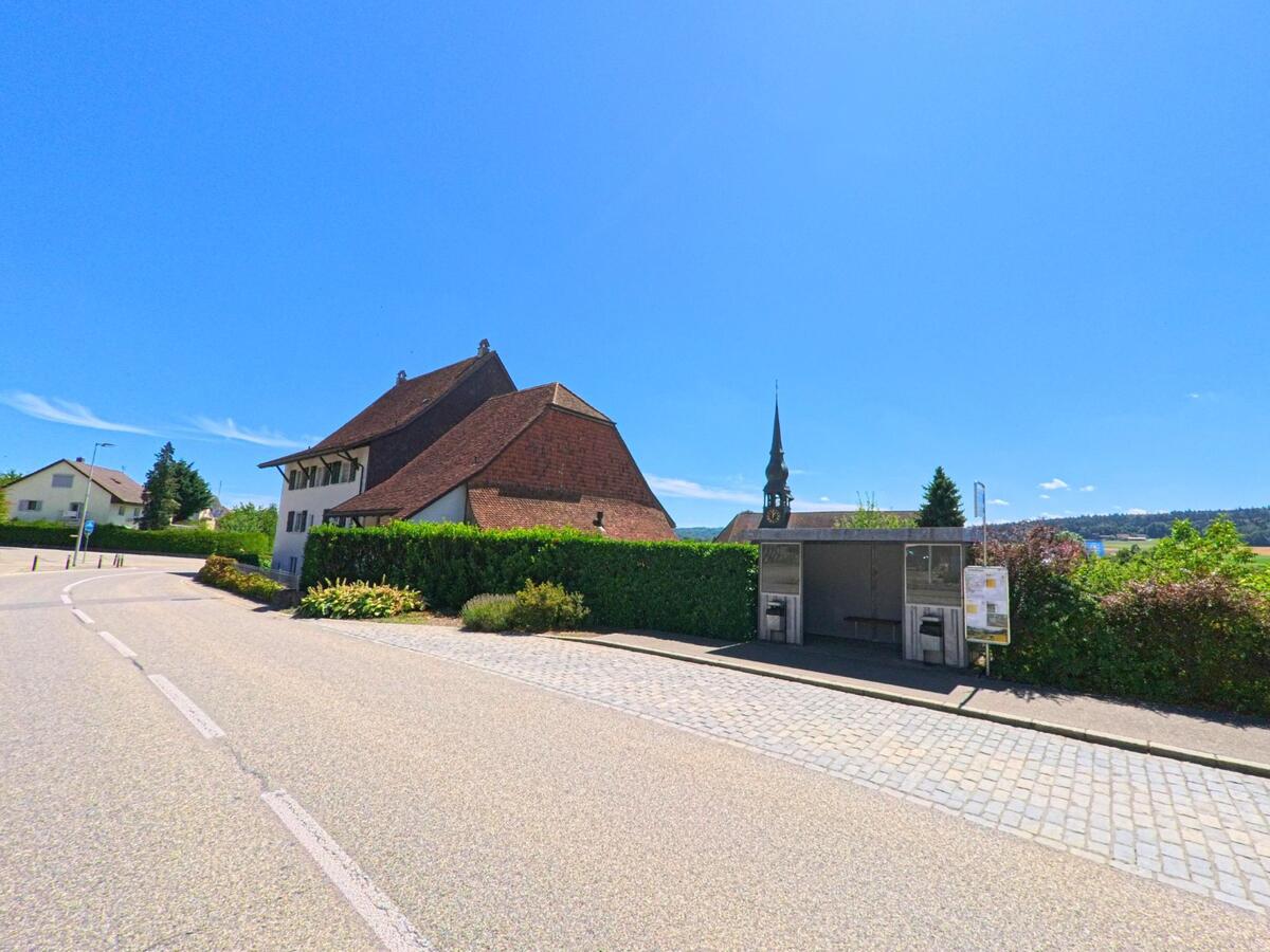 Eine überdachte Bushaltestelle am Strassenrand in Wolfwil (4628, Kanton Solothurn) bietet Blick auf die Kirche mit ihrem markanten Turm und die umgebende Landschaft.