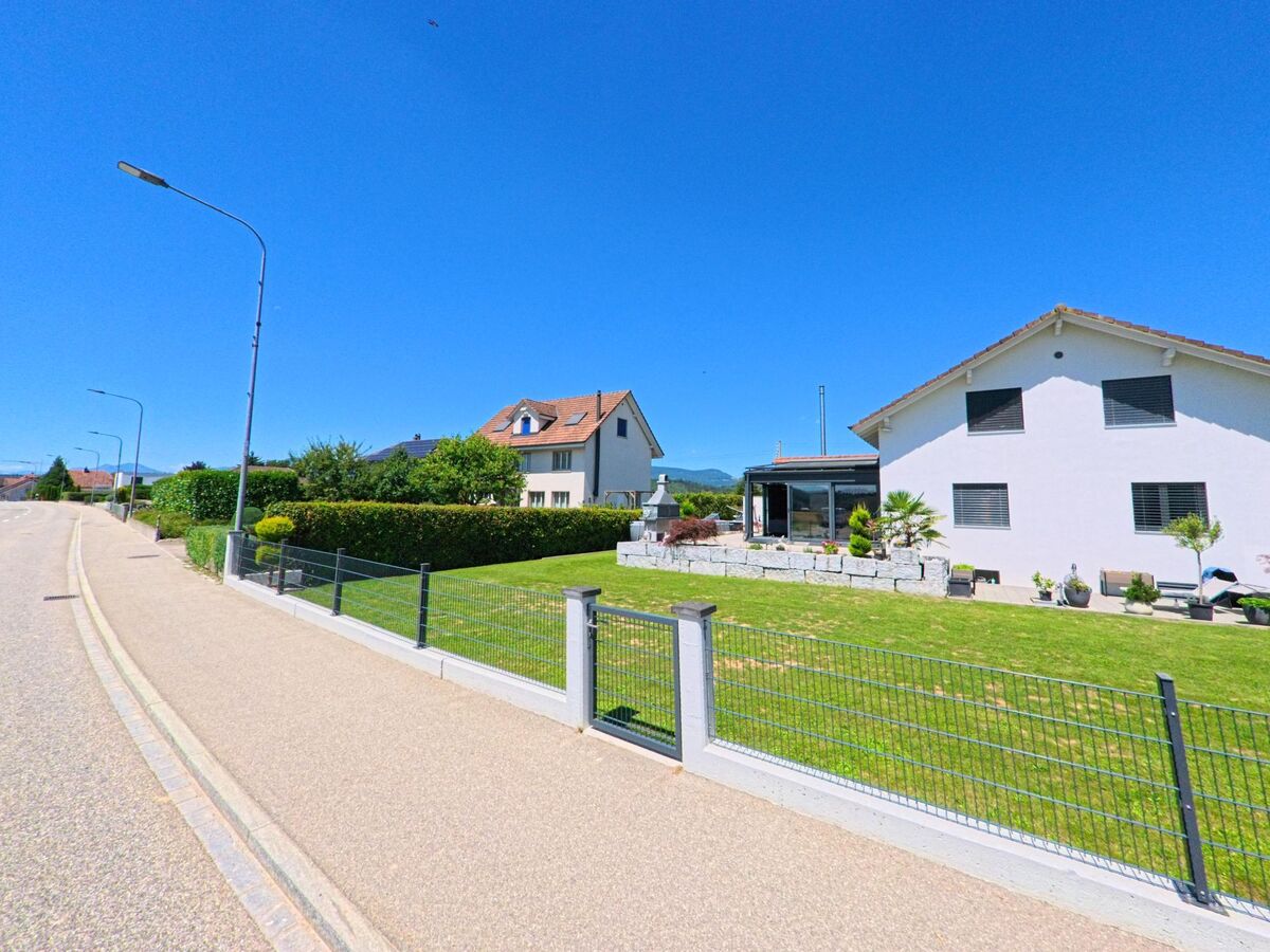 In Wolfwil (4628, Kanton Solothurn) säumen moderne Einfamilienhäuser mit gepflegten Vorgärten eine ruhige Strasse unter wolkenlosem Himmel.