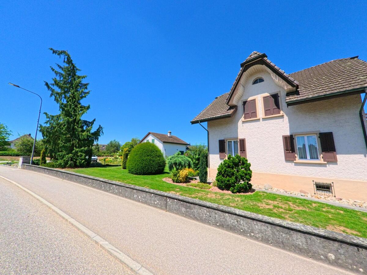 In Wolfwil (4628, Kanton Solothurn) steht ein gepflegtes Wohnhaus mit Fensterläden und vorgelagertem Garten an einer ruhig verlaufenden Strasse mit Laternen und Nadelbäumen.