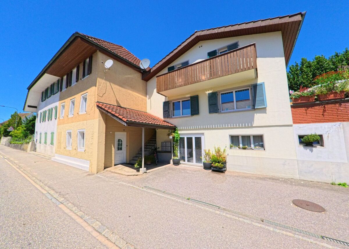 In Wolfwil (4628, Kanton Solothurn) reihen sich an einer schmalen Strasse Wohnhäuser mit Fensterläden, Balkonen und Blumenschmuck unter klarem Sommerhimmel.