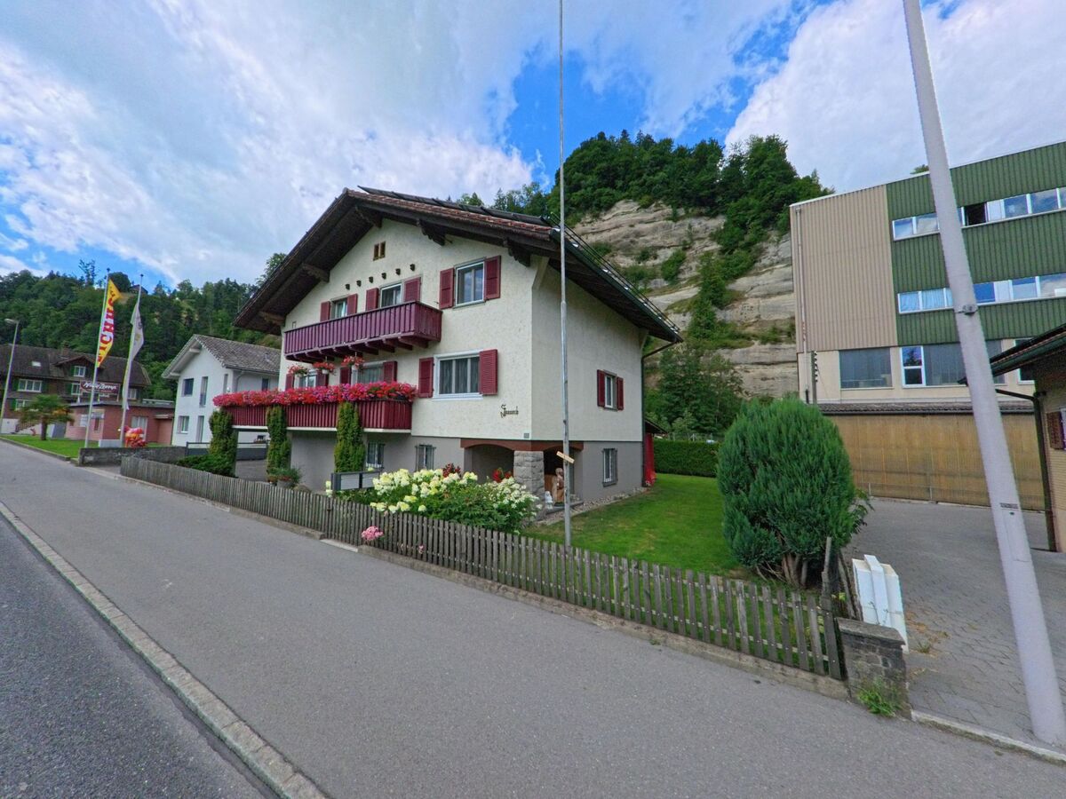  In Wolhusen (6110, Kanton Luzern) steht ein traditionelles Wohnhaus mit roten Fensterläden und Blumenschmuck vor einer markanten Felswand am Rand einer Wohnsiedlung.
