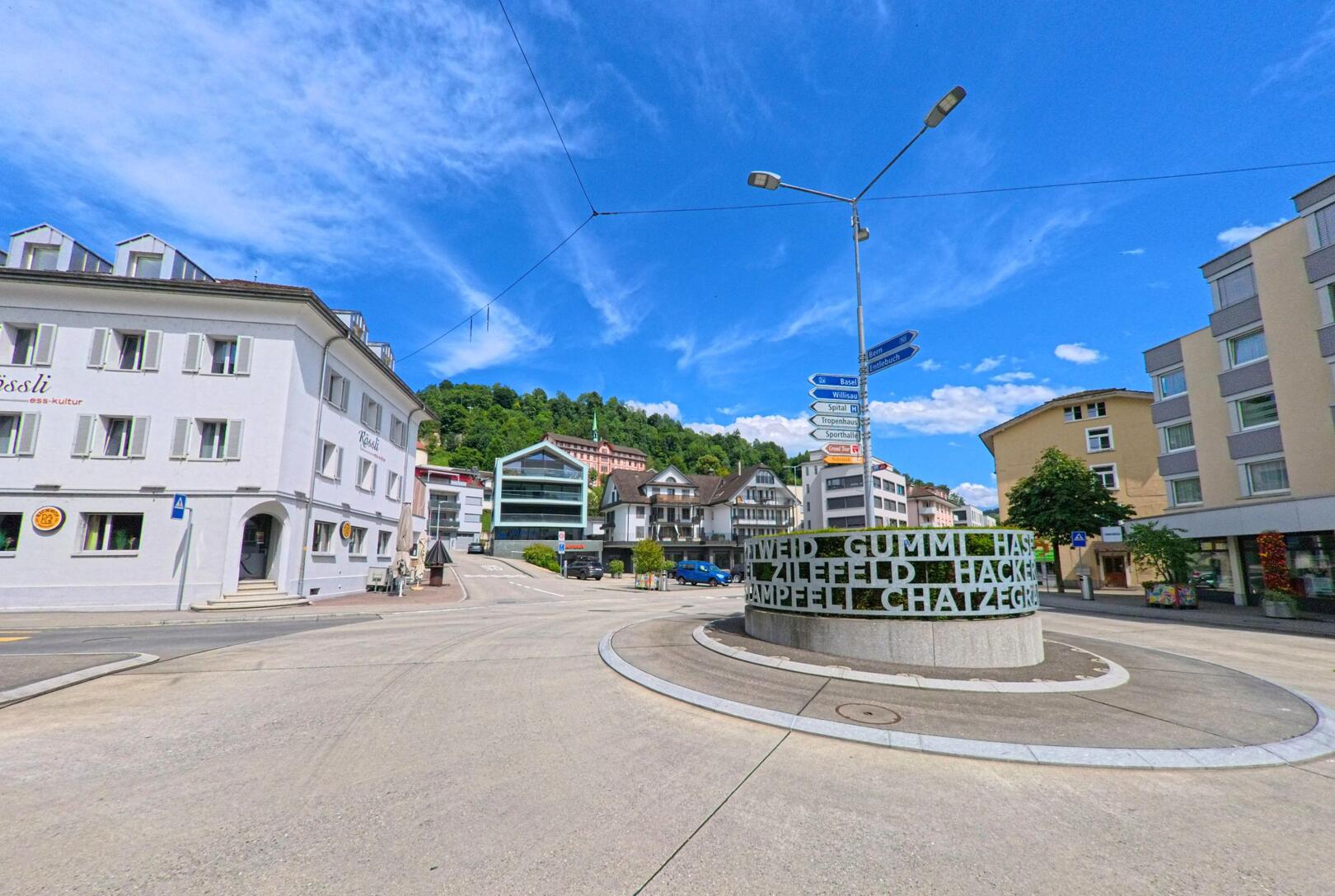 In Wolhusen (6110, Kanton Luzern) markiert ein moderner Kreisverkehr mit grün beschrifteter Skulptur den zentralen Treffpunkt zwischen Wohnhäusern und Geschäften.