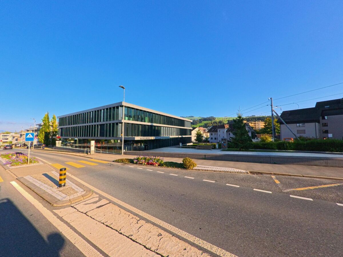 Vor dem modernen Gebaeude der Gemeinde Wollerau (8832, Kanton Schwyz) verlaeuft eine breite Strasse mit Mittelinsel und Zebrastreifen, eingerahmt von Blumenrabatten.