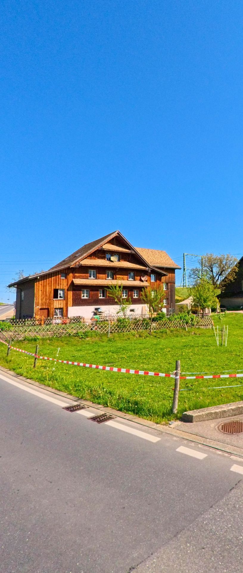 Ein traditionelles Holzhaus auf einer Wiese in Wollerau (8832, Kanton Schwyz) steht bei klarer Sicht und blauem Himmel am Rand einer Strasse.