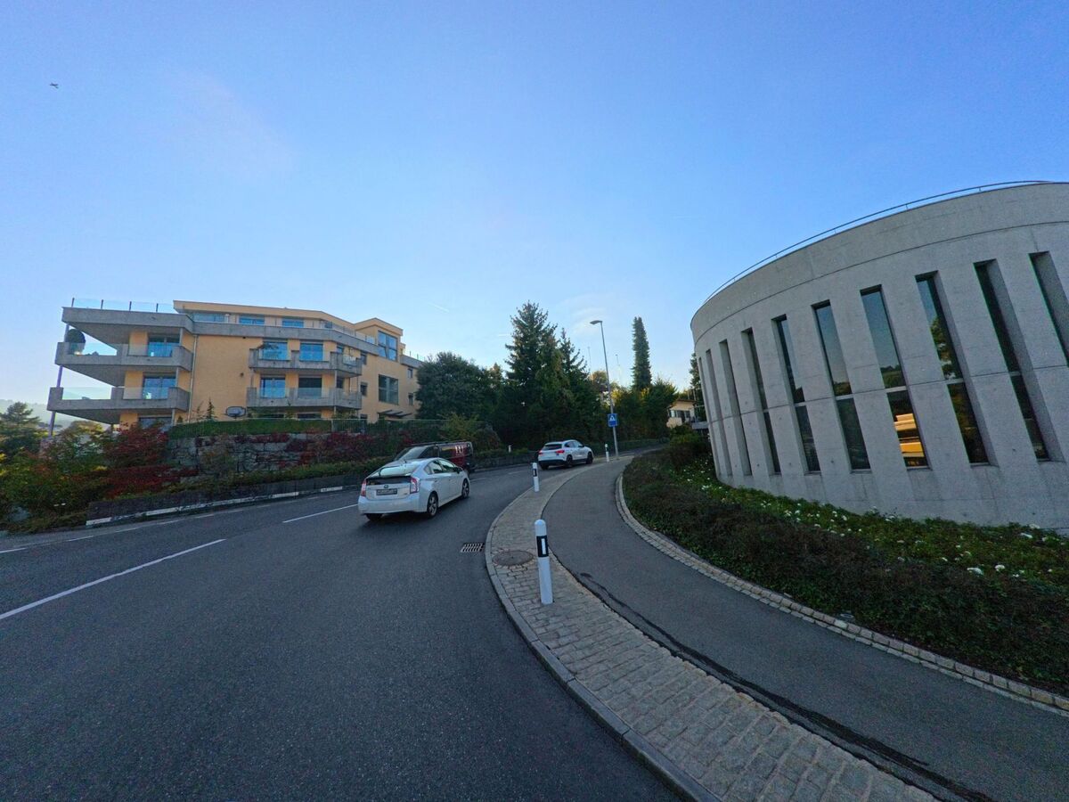 Eine leicht ansteigende Strasse in Wollerau (8832, Kanton Schwyz) fuehrt zwischen einem modernen Rundbau und einem mehrgeschossigen Wohnhaus hindurch.