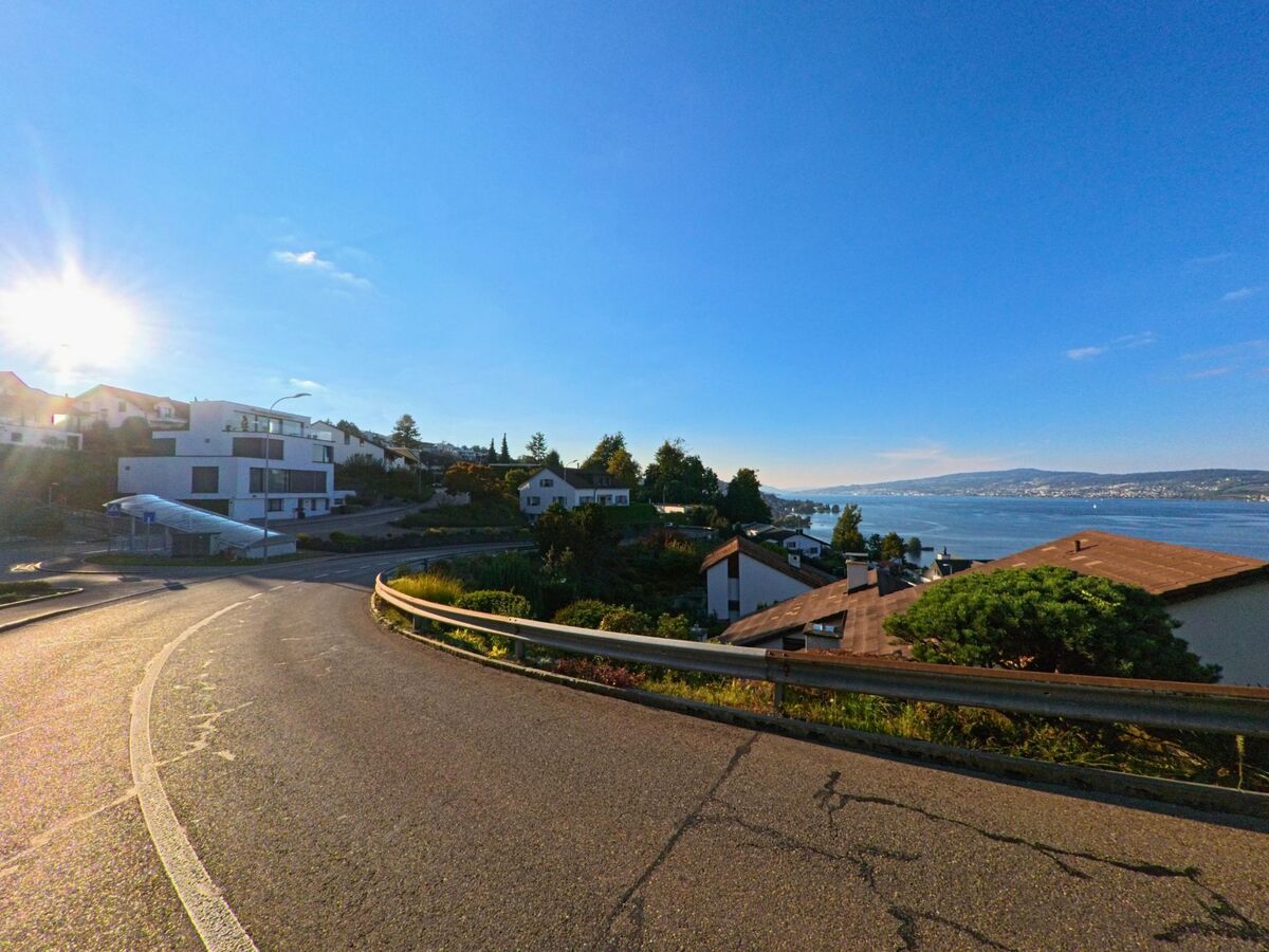 Entlang einer kurvigen Strasse in Wollerau (8832, Kanton Schwyz) oeffnet sich der Blick auf moderne Wohnhaeuser und die weite Wasserflaeche des Sees.