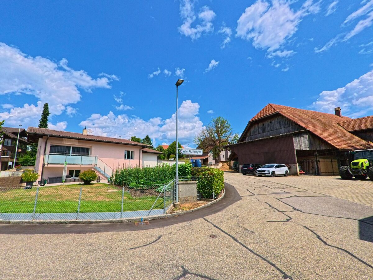 In einem Wohnquartier von Worb (3076, Kanton Bern) trifft ein moderner Bungalow auf ein traditionelles Bauernhaus mit Scheune und Traktor bei blauem Himmel.