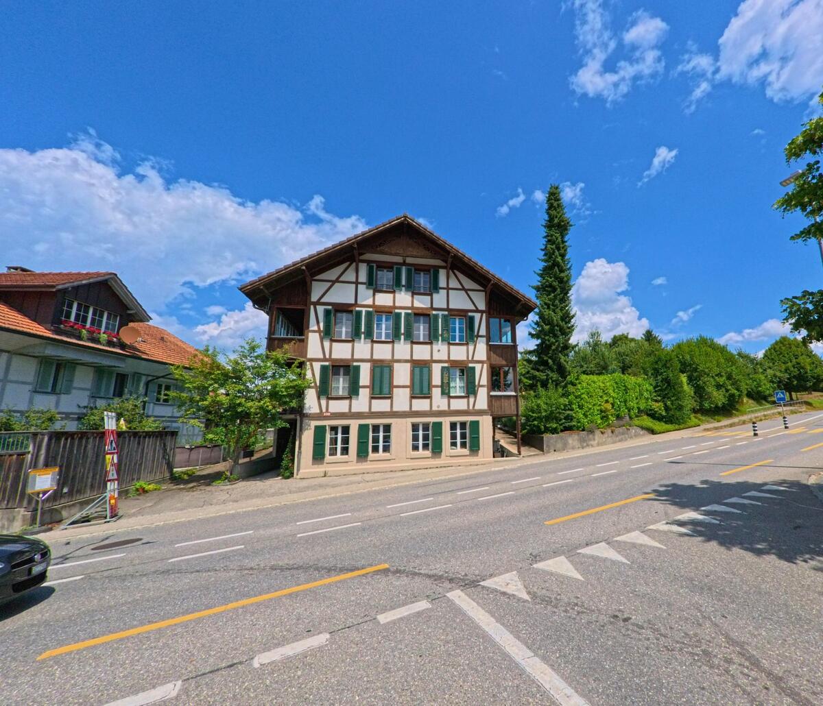 Direkt an einer Hauptstrasse in Worb (3076, Kanton Bern) steht ein stattliches Fachwerkhaus mit gruenen Fensterlaeden und gepflegtem Vorgarten.