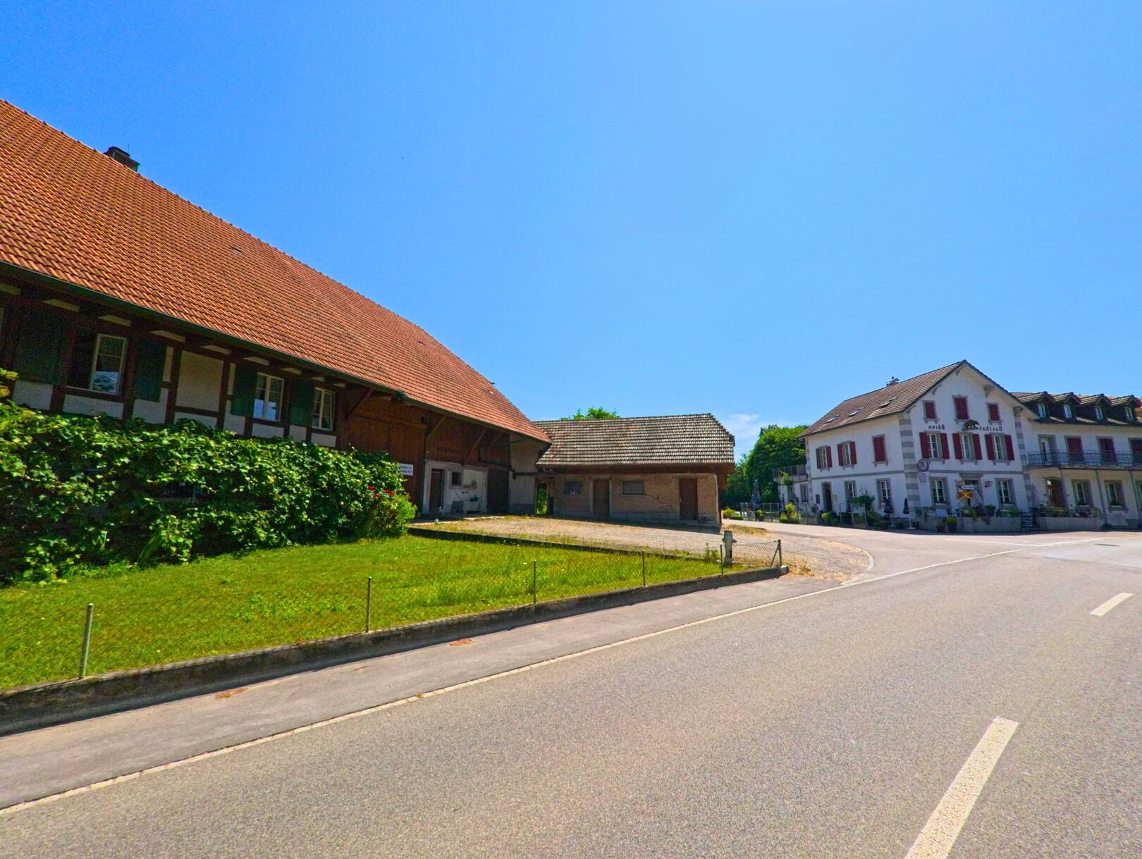 In Worben (3252, Kanton Bern) verläuft eine Strasse entlang eines traditionellen Fachwerkhauses und eines modernen Gebäudes, umgeben von grünen Wiesen und Bäumen.