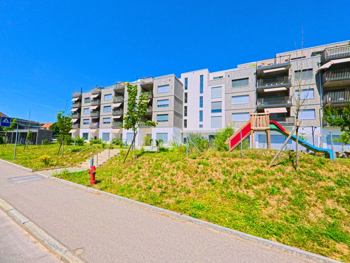 In Worben (3252, Kanton Bern) grenzen moderne Mehrfamilienhäuser mit Balkonen an einen Spielplatz mit Rutsche entlang eines Gehwegs mit Zebrastreifen.
