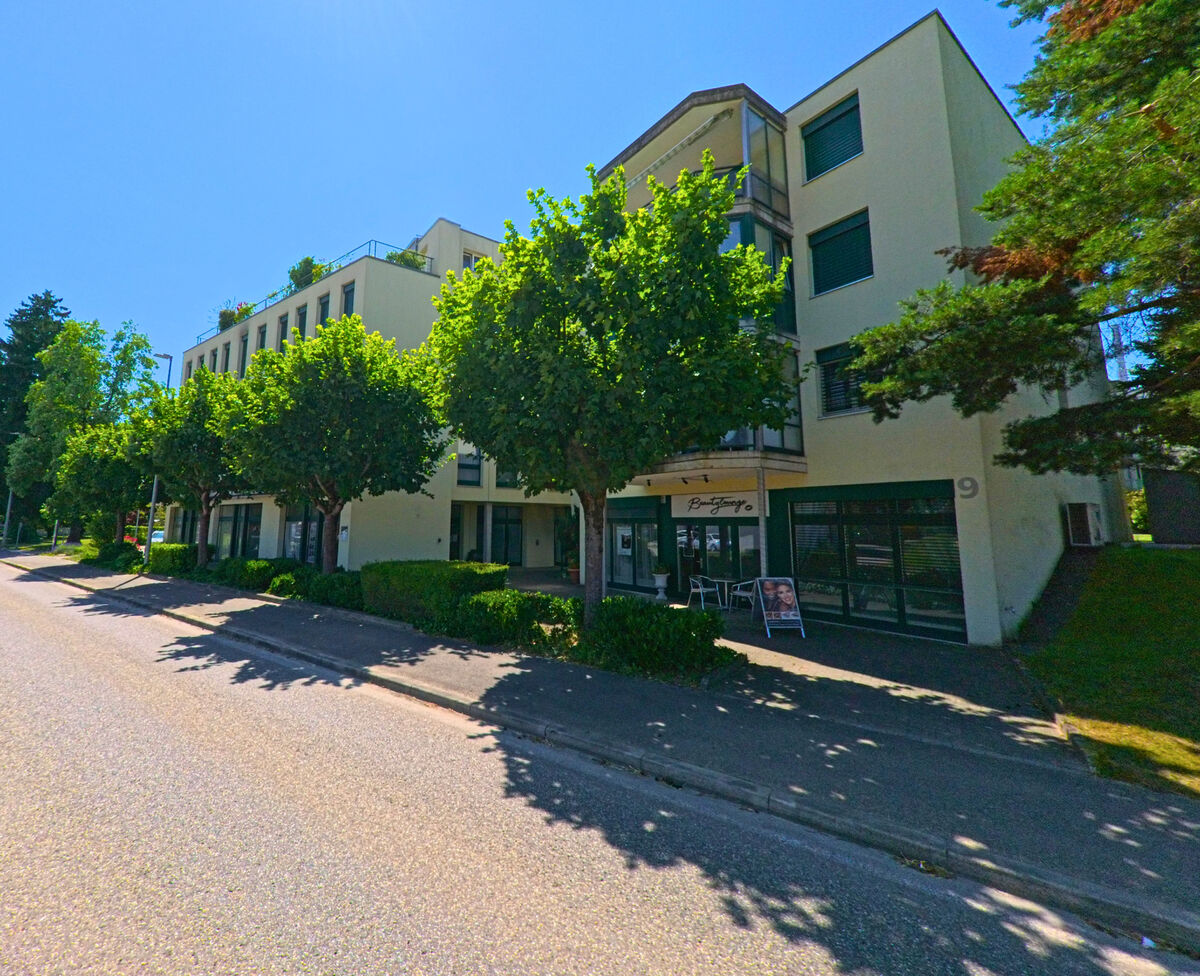 In Worben (3252, Kanton Bern) steht ein modernes Wohn- und Geschäftsgebäude mit schattenspendenden Bäumen entlang einer breiten Strasse.