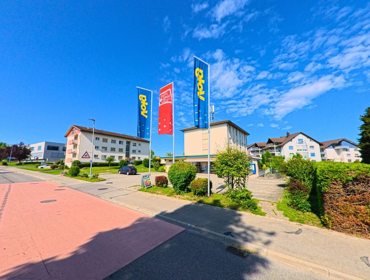 Vor einem Geschäft in Wünnewil-Flamatt (3184, Kanton Freiburg) wehen mehrere Fahnen im Wind, umgeben von Wohnhäusern und einer ruhigen Strasse.