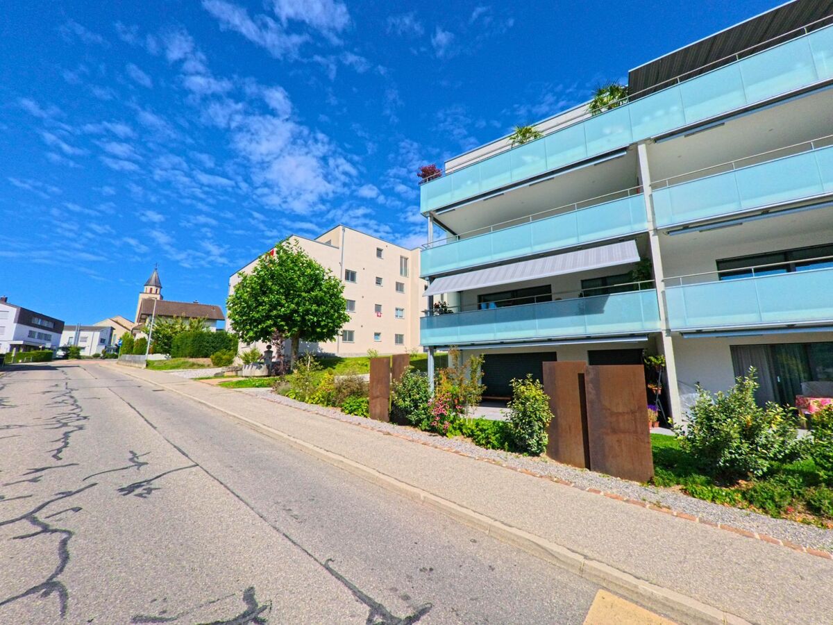 Moderne Wohnbauten in Wünnewil-Flamatt (3184, Kanton Freiburg) stehen entlang einer ansteigenden Strasse, im Hintergrund ist der Kirchturm der Kirche zu sehen.