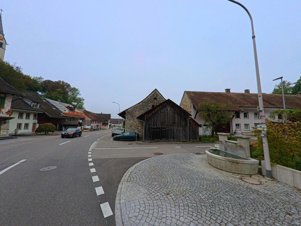 Ein gepflasterter Platz mit Brunnen in Wuerenlingen (5303, Kanton Aargau) liegt an einer sanft geschwungenen Strasse, flankiert von alten Wohn- und Scheunenfassaden.