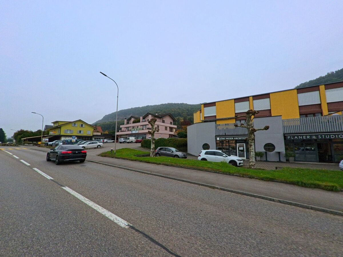 Mehrere Wohnhaeuser und Gewerbeflaechen saeumen in Wuerenlingen (5303, Kanton Aargau) eine breite Strasse vor dem bewaldeten Hang.
