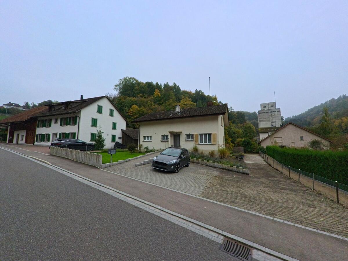 Zwei Wohnhaeuser am Hang von Wuerenlingen (5303, Kanton Aargau) liegen an einer ruhigen Strasse, waehrend ein Industriebau im Talbereich sichtbar ist.