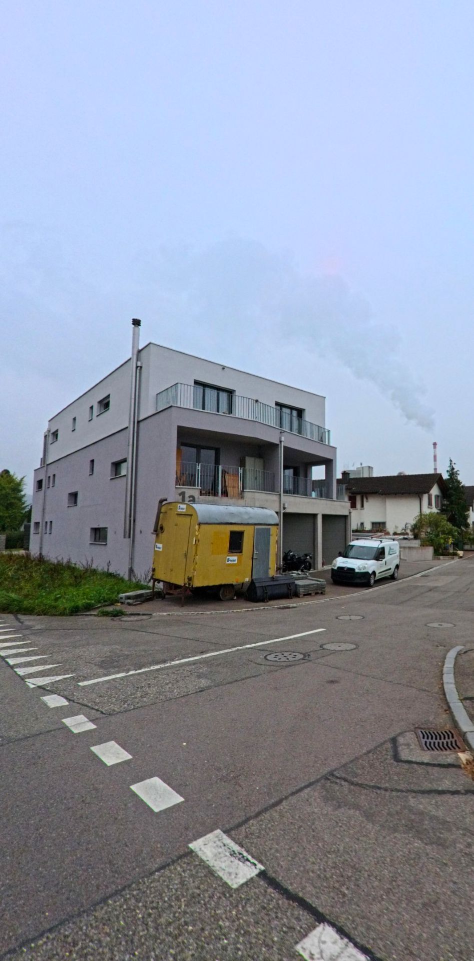 Ein modernes Wohnhaus in Wuerenlingen (5303, Kanton Aargau) steht an einer ruhigen Kreuzung, waehrend im Hintergrund Rauch aus einer Industrieanlage aufsteigt.