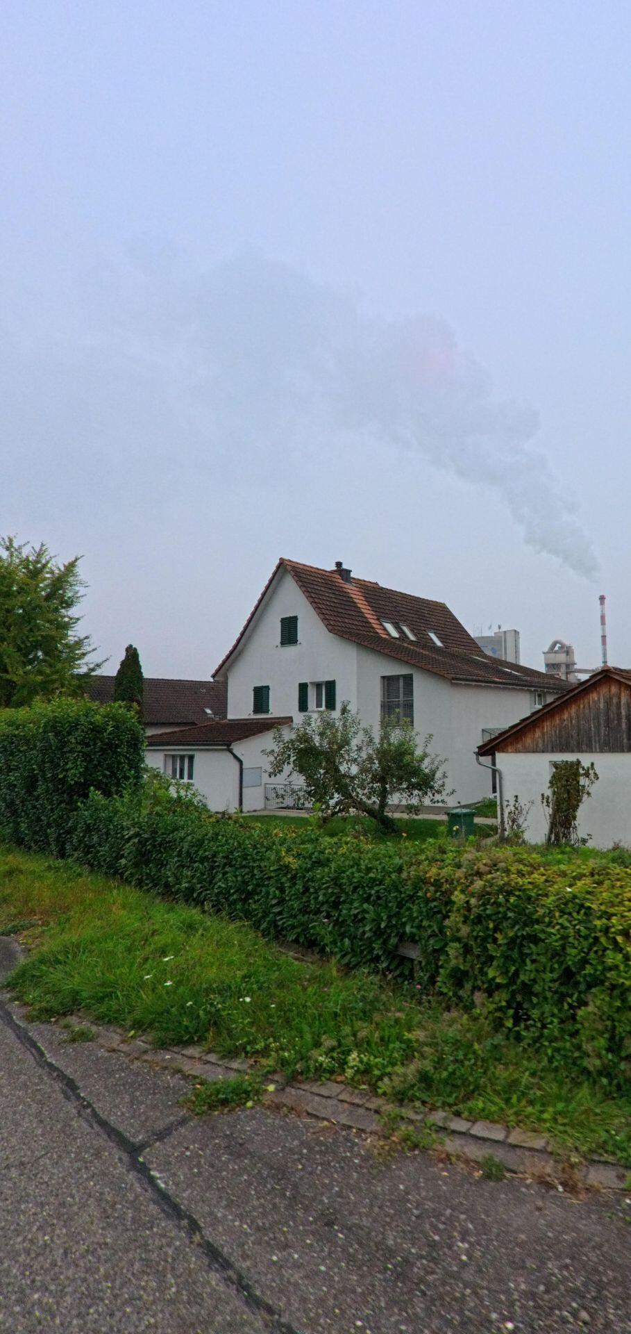 Hinter einem Wohnhaus in Wuerenlingen (5303, Kanton Aargau) steigt dichter Rauch aus einer Industrieanlage auf, waehrend eine Hecke den Gehweg saeumt.