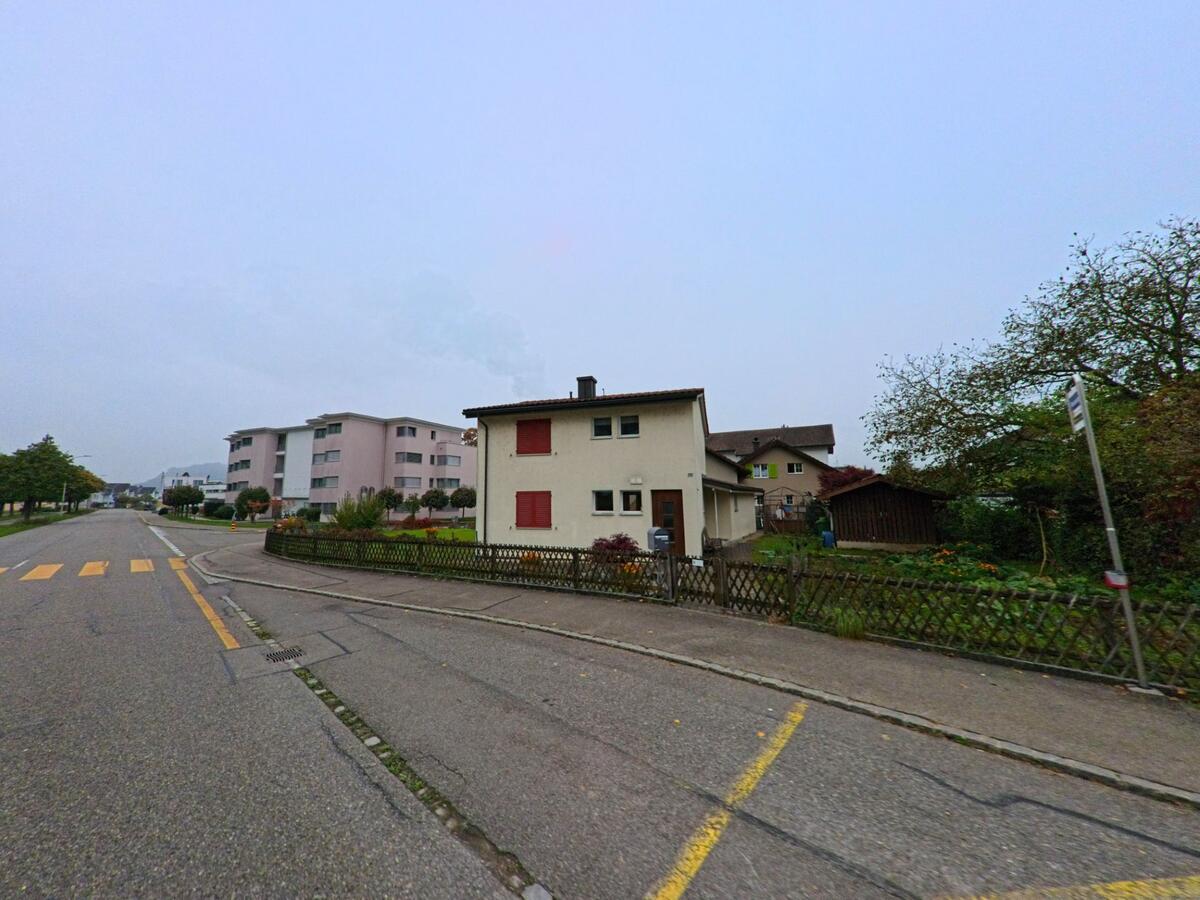 Ein kleines Wohnhaus mit Garten liegt in Wuerenlingen (5303, Kanton Aargau) an einer breiten Strasse, waehrend moderne Wohnbloecke die Gegend praegen.