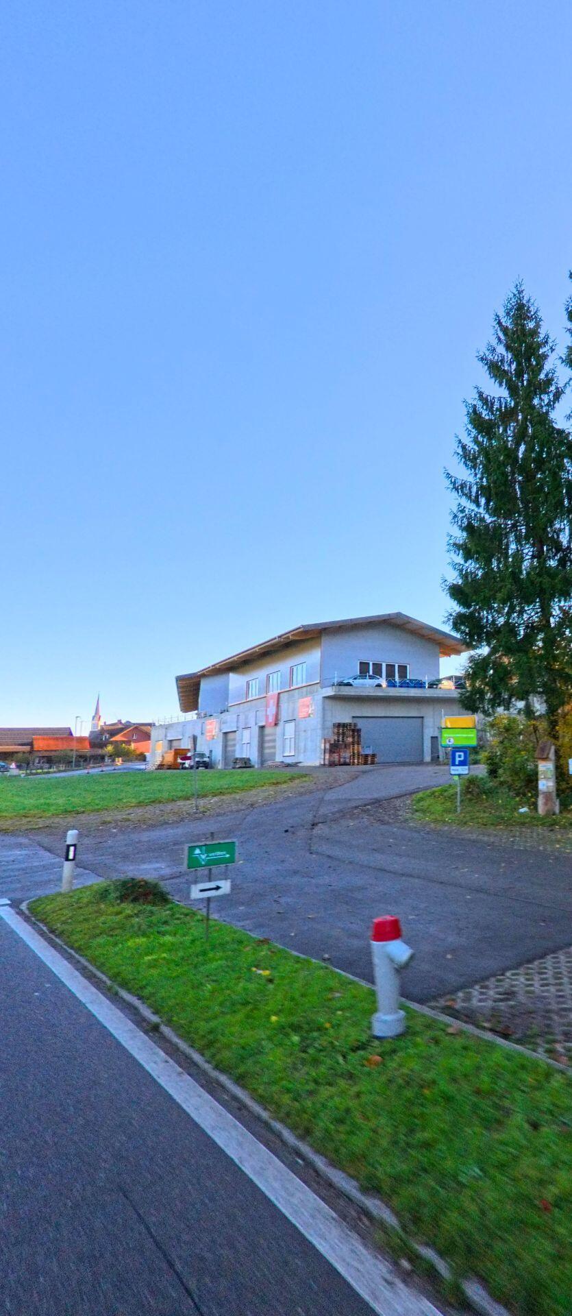 An der Zufahrt zu einem Gewerbegebaeude in Wuppenau (9514, Kanton Thurgau) fuehrt eine asphaltierte Strasse an Gruenflaechen und Wegweisern vorbei unter klarem Himmel.