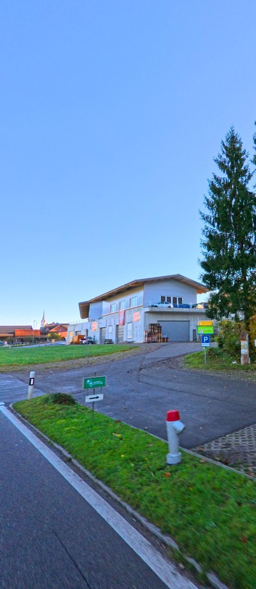 An der Zufahrt zu einem Gewerbegebaeude in Wuppenau (9514, Kanton Thurgau) fuehrt eine asphaltierte Strasse an Gruenflaechen und Wegweisern vorbei unter klarem Himmel.