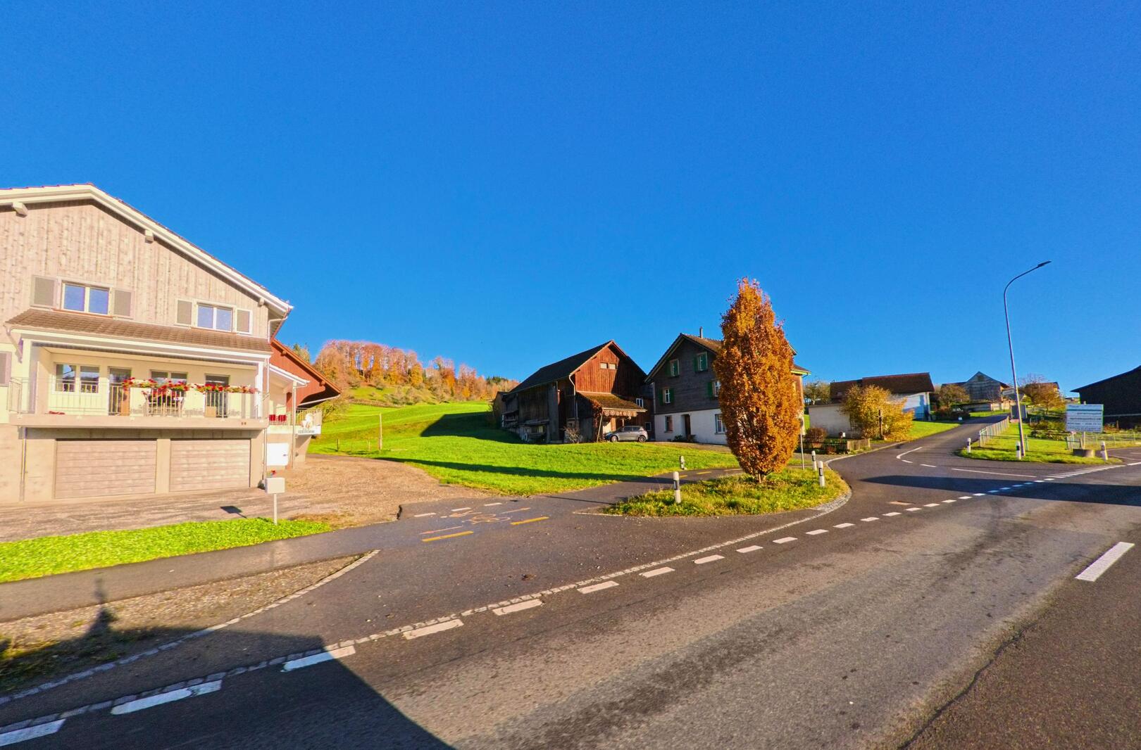 An einer ruhigen Strassenkreuzung in Wuppenau (9514, Kanton Thurgau) stehen ländliche Wohnhaeuser neben gruenen Wiesen und herbstlich gefaerbten Baeumen unter klarem Himmel.