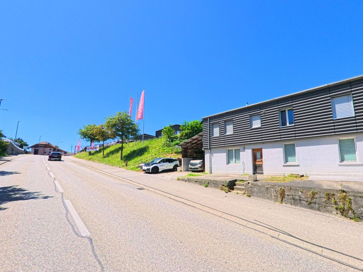 Entlang einer ansteigenden Strasse in Wynau (4923, Kanton Bern) befinden sich moderne Gebaeude und Parkplaetze mit Werbeflaggen auf einer kleinen Anhöhe.