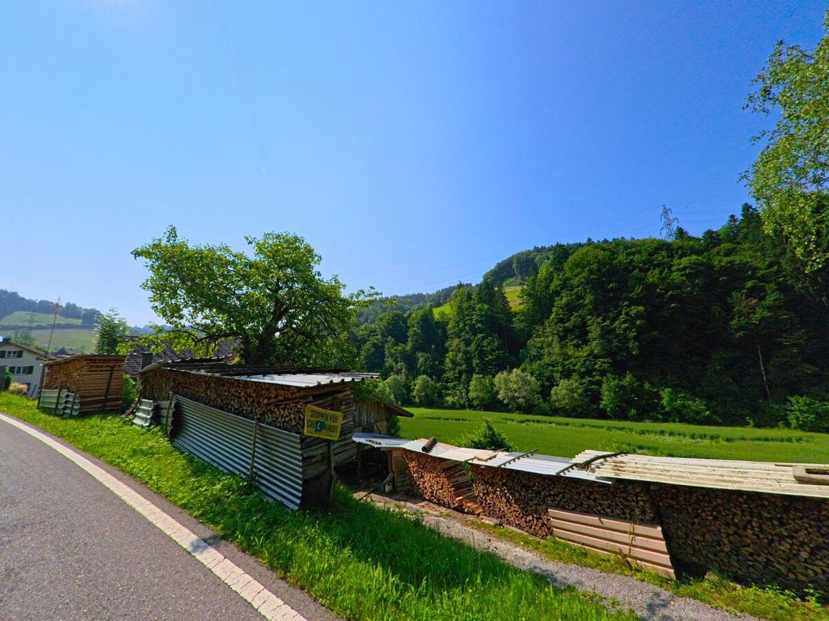 In Wynigen (3472, Kanton Bern) lagert Brennholz ordentlich gestapelt entlang einer kurvigen Landstrasse mit Blick auf ein bewaldetes Tal.
