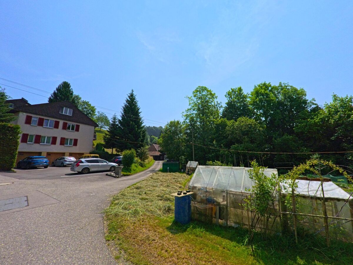 Am Rand eines Wohnquartiers in Wyssachen (4954, Kanton Bern) stehen mehrere Gewaechshaeuser neben einem Weg, der zwischen Waldrand und Haeusern verlaeuft.