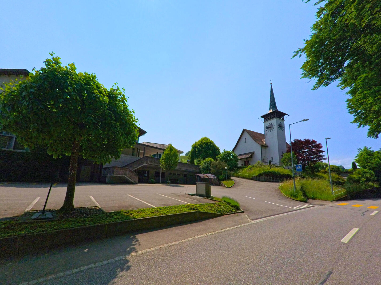 Auf einer Anhöhe in Wyssachen (4954, Kanton Bern) erhebt sich eine Kirche mit spitzem Turm über einem Parkplatz und einer leicht ansteigenden Strasse.