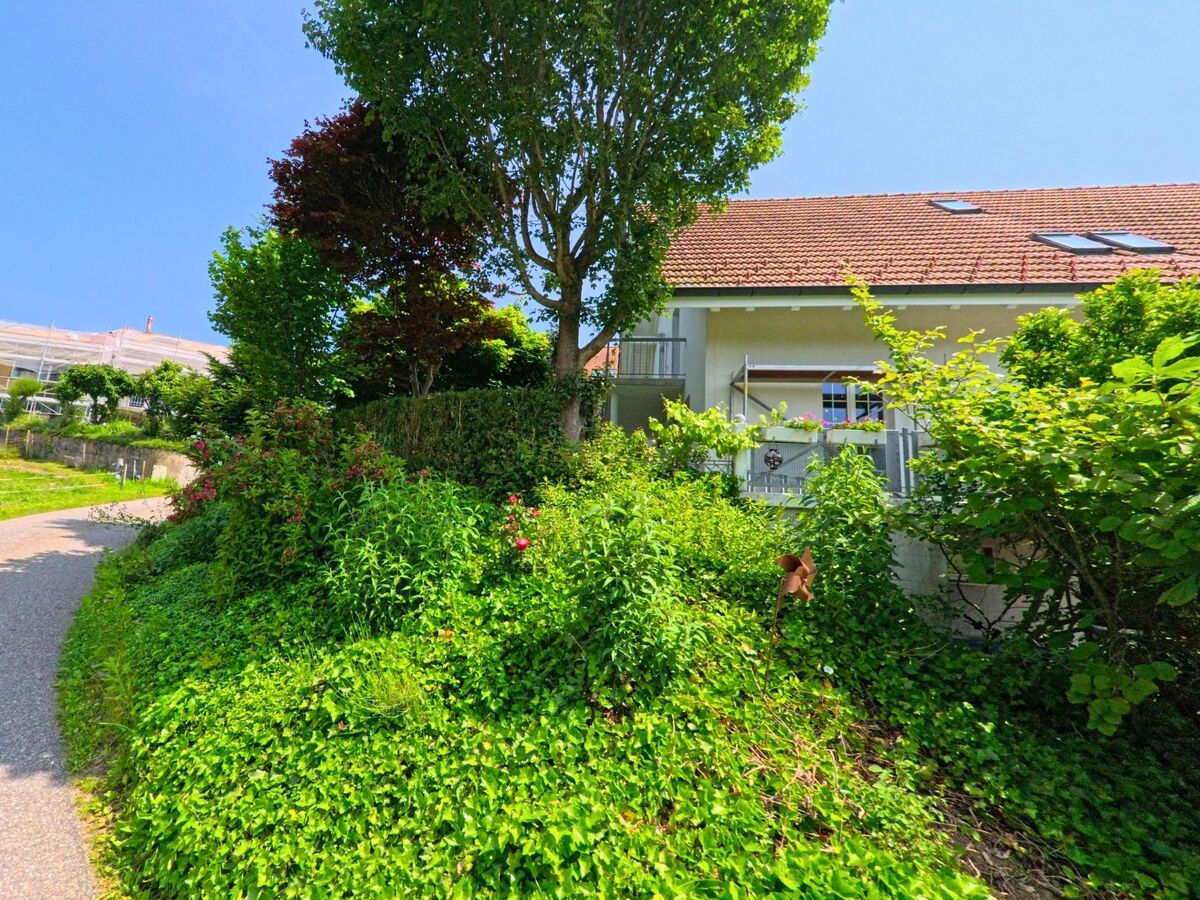 In Wyssachen (4954, Kanton Bern) liegt ein Wohnhaus mit rotem Ziegeldach leicht oberhalb eines Weges, umgeben von dichter Bepflanzung und Blueten.