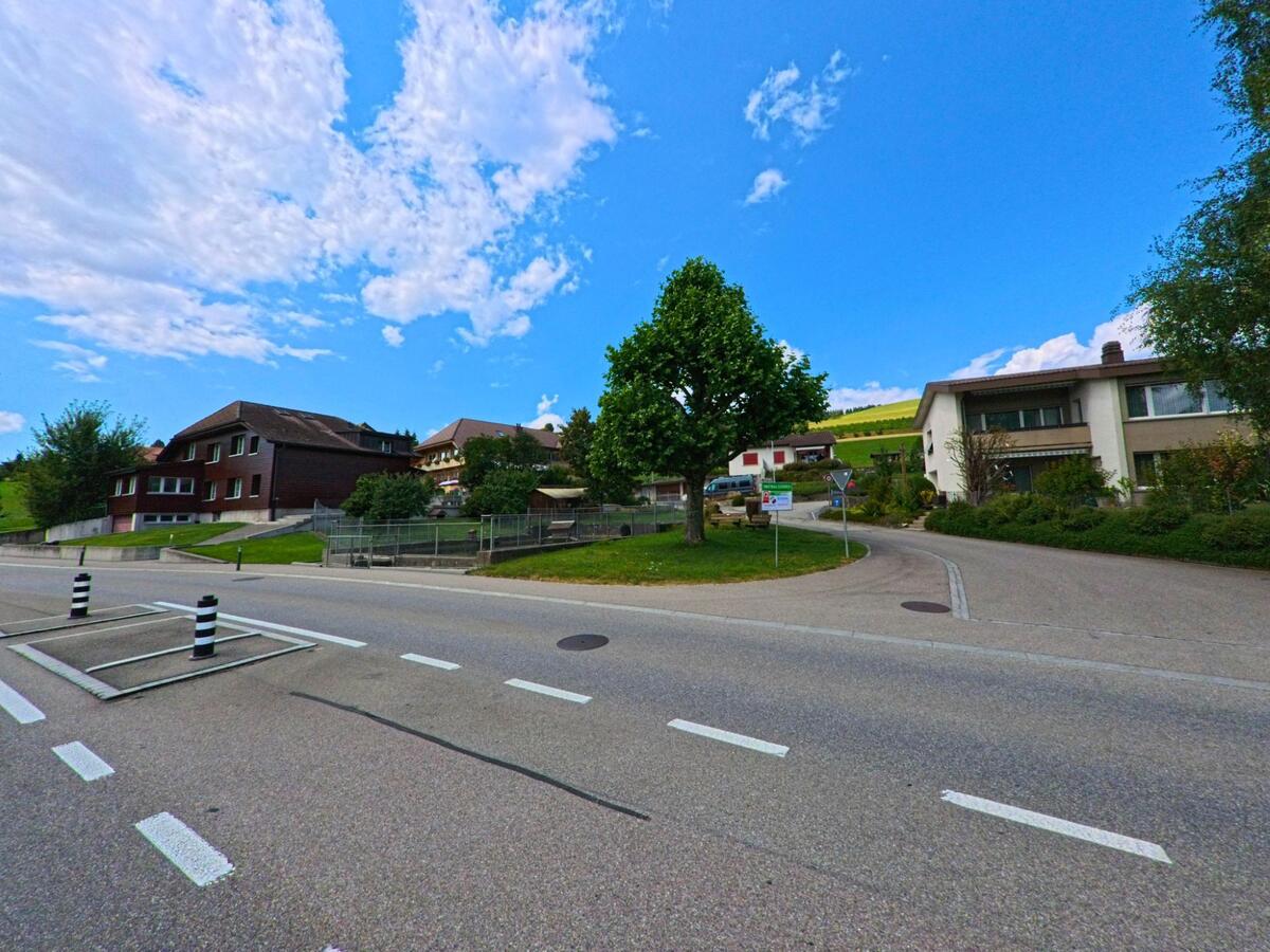 Am Rand von Zaeziwil (3532, Kanton Bern) zweigt eine Nebenstrasse mit Baumbestand und Wohnhaeusern in ein ruhiges Quartier mit Blick auf die huegelige Landschaft ab.