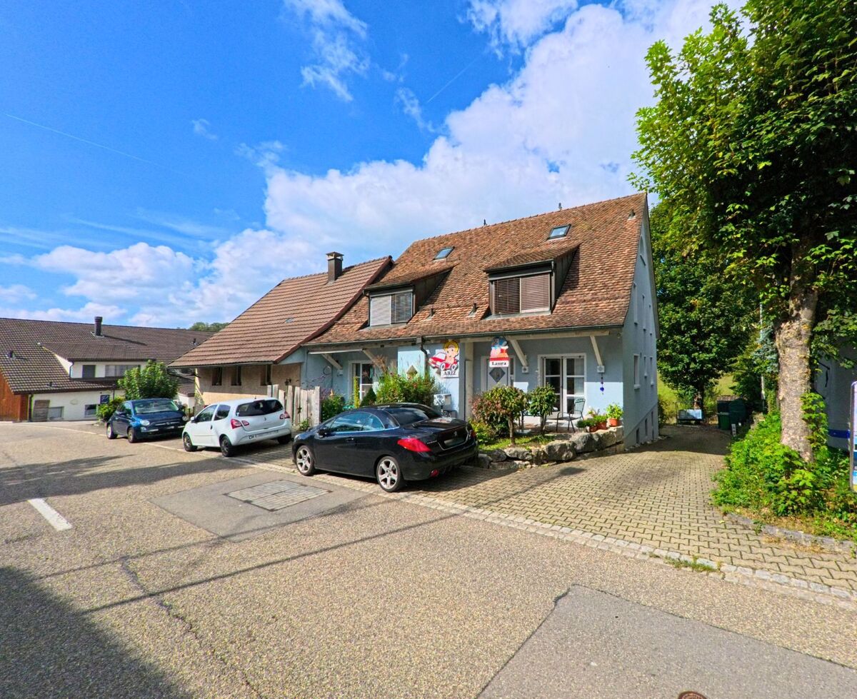 In Zeihen (5079, Kanton Aargau) stehen zwei Wohnhaeuser mit Vorgarten und parkenden Autos, waehrend ein Baum Schatten auf die Einfahrt wirft.