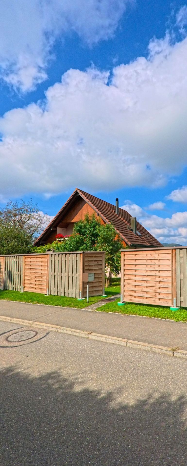 In Zeihen (5079, Kanton Aargau) steht ein Haus mit steilem Satteldach hinter hohen Holzabschirmungen, umgeben von dichtem Gruen und einzelnen Blueten.