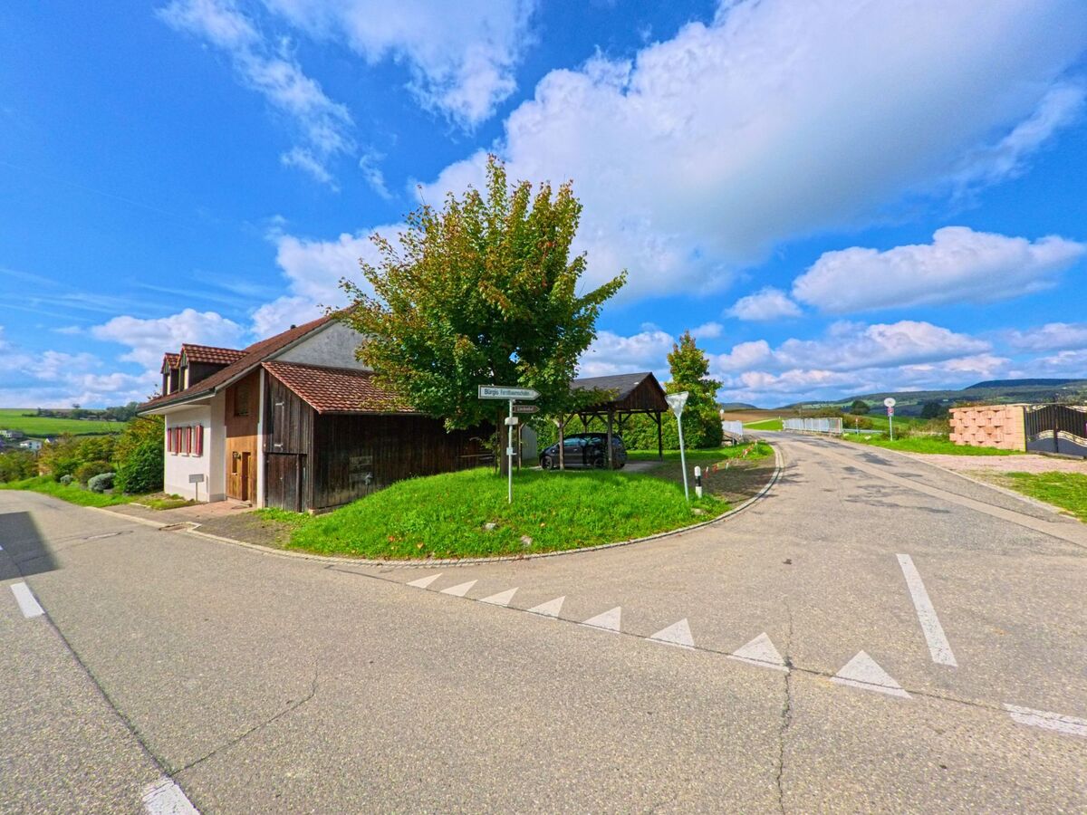 In Zeihen (5079, Kanton Aargau) treffen zwei schmale Strassen aufeinander, neben einem alten Bauernhaus und einer kleinen ueberschatteten Parkflaeche.