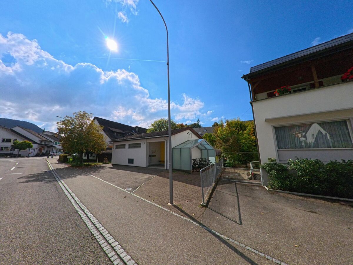 In Zeihen (5079, Kanton Aargau) stehen helle Wohnhaeuser entlang einer breiten Strasse, waehrend ein kleiner Fahrradunterstand neben Bueschen liegt.