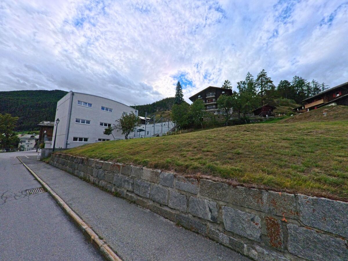 Ein modernes Gebaeude und mehrere Wohnhaeuser in Zeneggen (3934, Kanton Wallis) liegen am Hang oberhalb einer Steinmauer mit Gehweg.