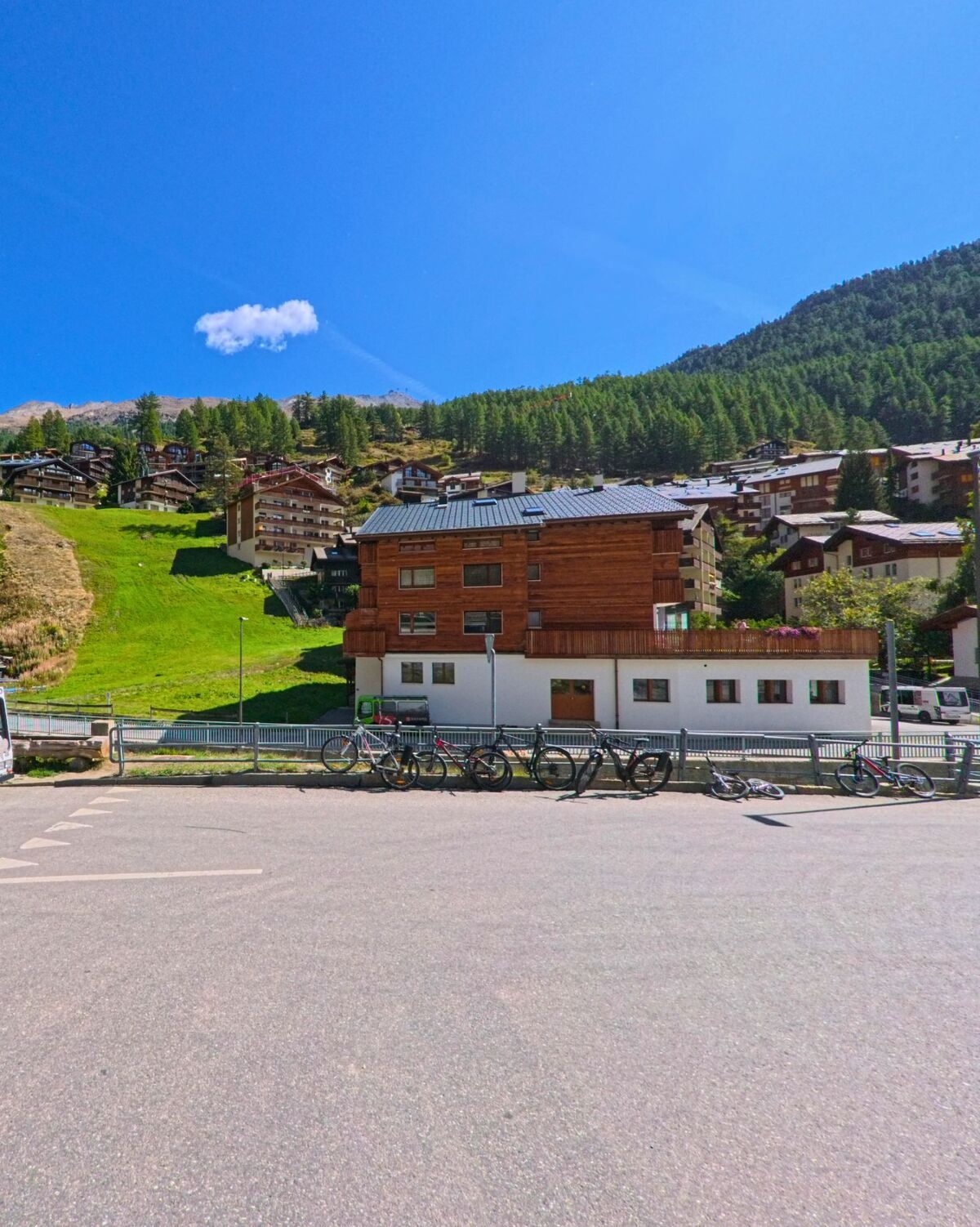 Vor einem modernen Holzhaus in Zermatt (3920, Kanton Wallis) sind mehrere Fahrraeder abgestellt, im Hintergrund liegen Haeuser am bewaldeten Berghang.