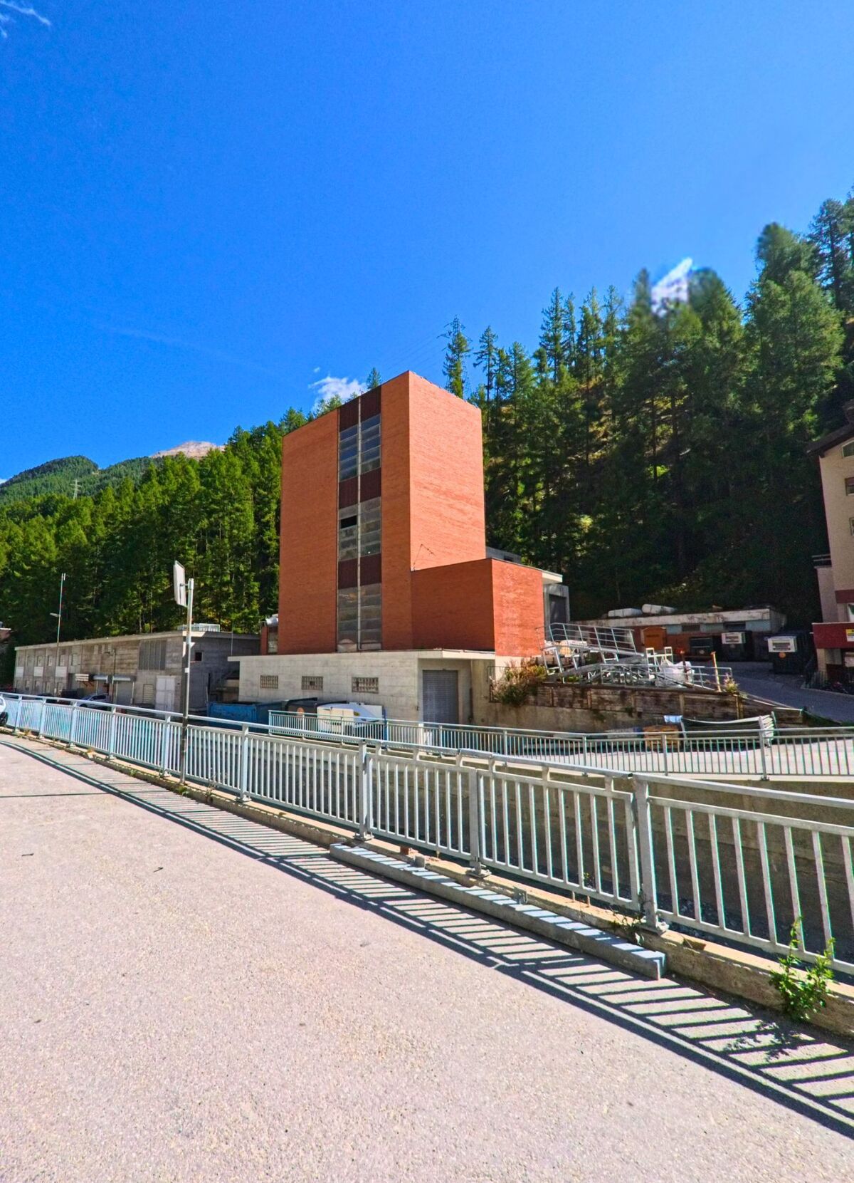 Ein roter, rechteckiger Turmbau steht in Zermatt (3920, Kanton Wallis) vor einem bewaldeten Hang und ist von einem Metallgelaender zur Strasse abgegrenzt.